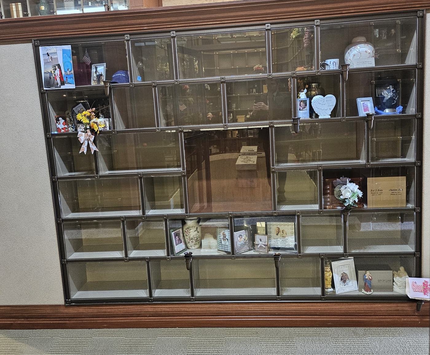 A face of the Eternal Light Columbarium with many empty glass-front niches of several sizes, a few of them holding urns, photographs and keepsakes