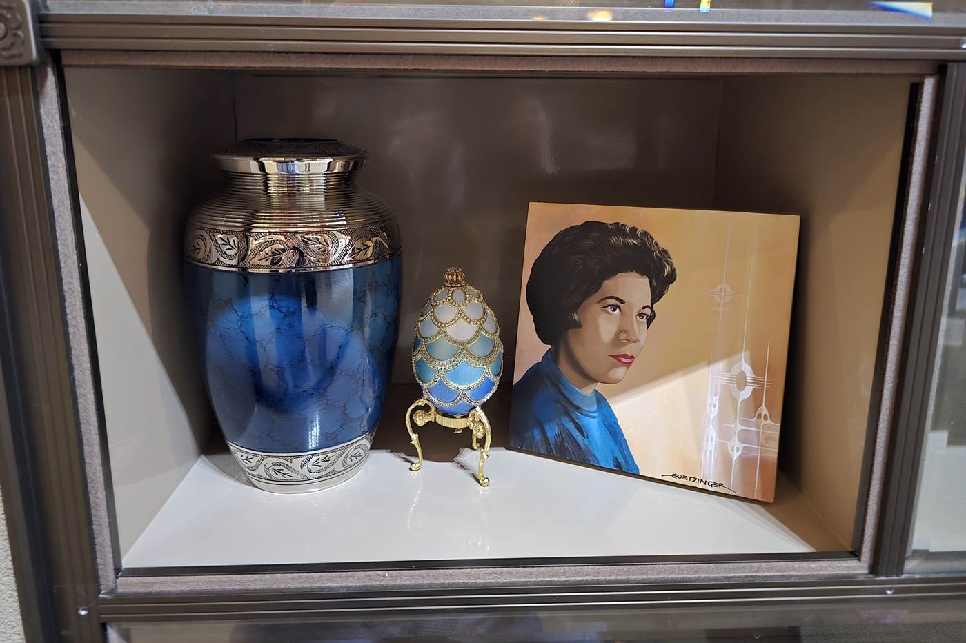 Close view inside one glass-front niche: a blue and silver urn, a jewelled keepsake egg on a gold stand, and a painted portrait of the woman interred, on a white shelf behind a single pane of glass