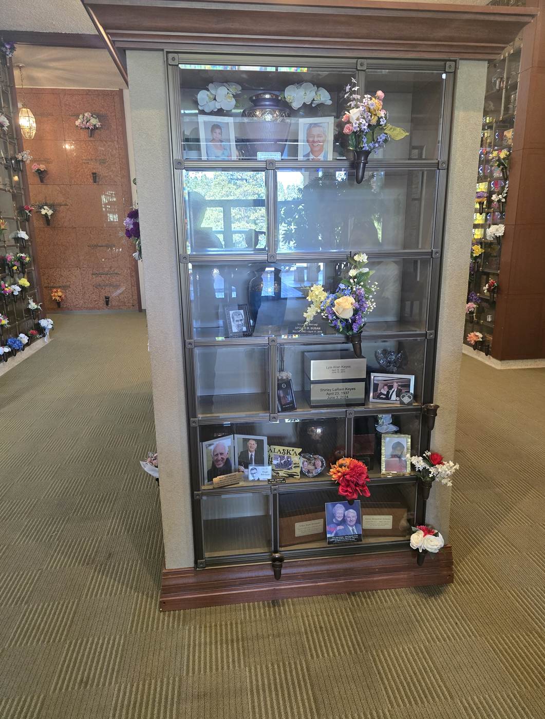 A four-sided glass-front columbarium at the Eternal Light Columbarium: urns, framed photographs and keepsakes visible through the glass, with bronze vases holding flowers on the frames