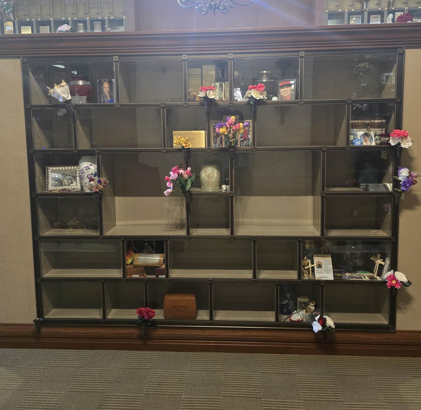 A built-in glass niche wall at the Eternal Light Columbarium, seen square on: rows of bronze-framed glass compartments in several sizes, some holding urns, framed pictures and flowers, many still empty