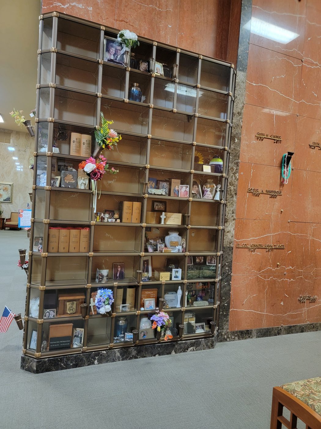 The Serenity niche wall: a bronze-framed glass niche wall against marble, with urns, books, framed pictures and flowers inside, and lettered crypt fronts on the wall alongside
