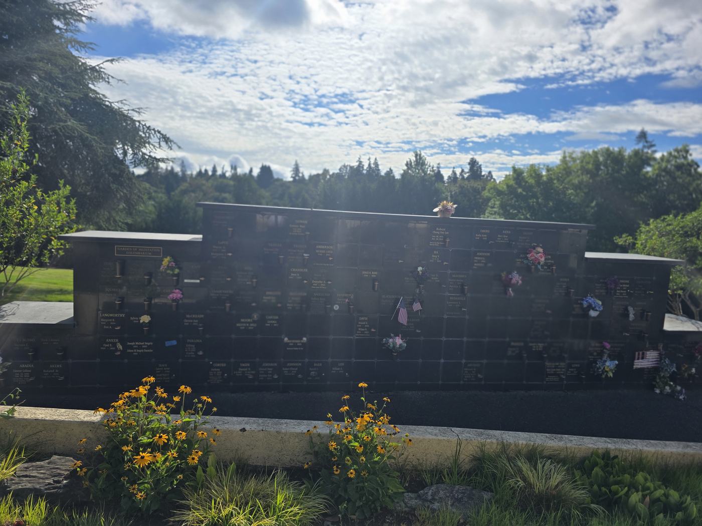 The Garden of Meditation columbarium wall in morning light: a long wall of polished black granite with bronze name plates and bronze vase holders, stepping up from low wings at either end to a taller centre section, with black-eyed Susans flowering in the bed in front of it