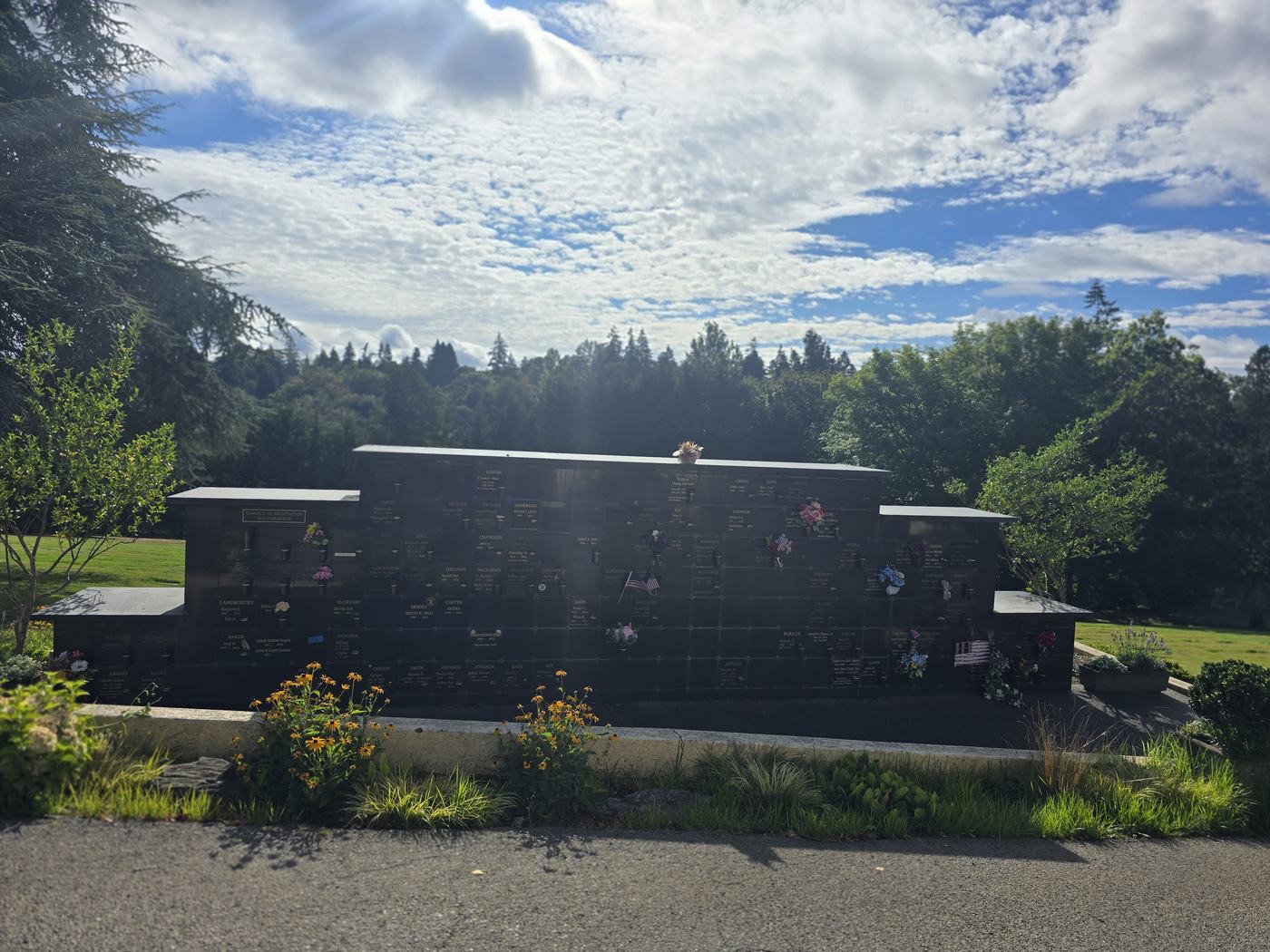 The Garden of Meditation wall seen from the roadway: the full stepped shape with its low wings, taller centre section, bronze plates and flowers left at several niches, under a partly cloudy sky