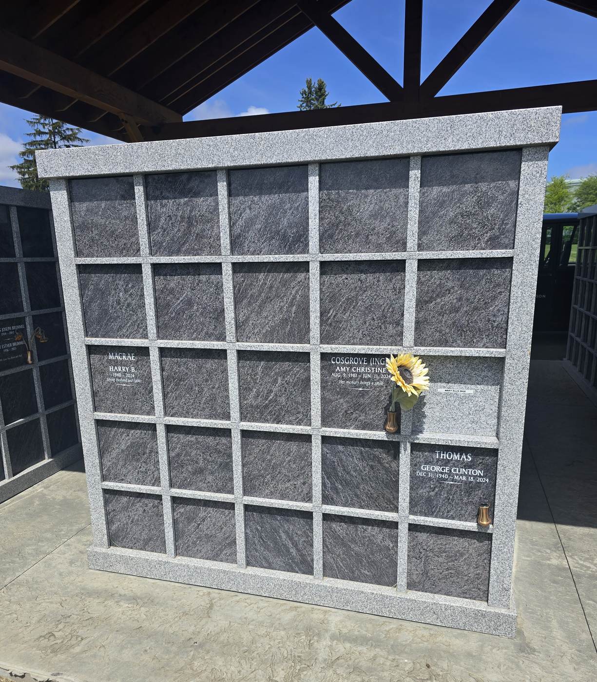 A Rock of Ages niche wall square on: a five-by-five grid of dark granite fronts in a light granite frame, three of them engraved with names and dates, one holding a bronze vase with a sunflower