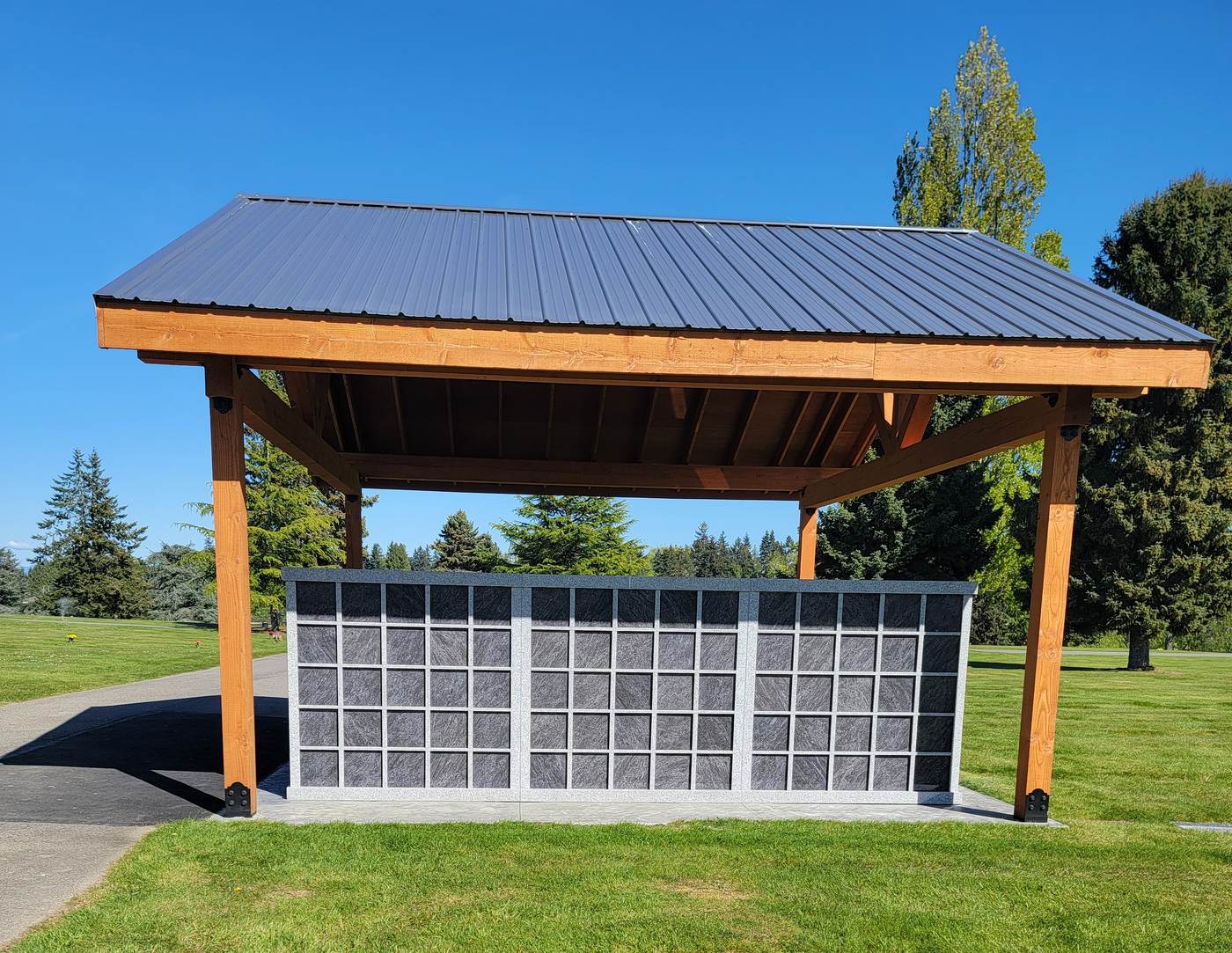 The Rock of Ages Columbarium from the lawn: a long granite niche wall of five tiers standing under an open timber pavilion with a dark metal roof, against a clear blue sky