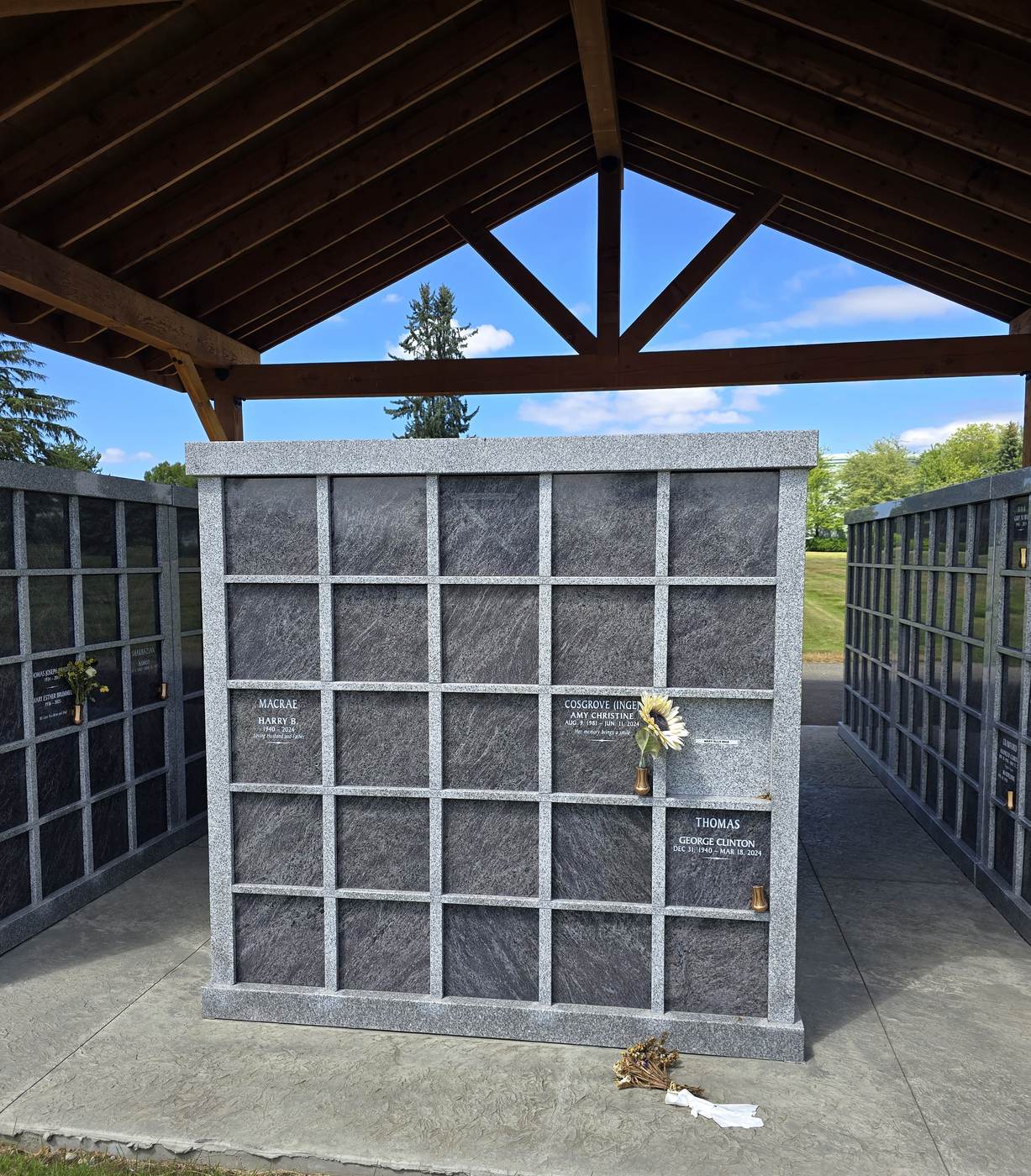 A granite niche wall at the Rock of Ages Columbarium under its timber pavilion: a five-by-five grid of dark granite niche fronts in a light granite frame, three of them engraved with names and dates, with a bronze vase holding a sunflower
