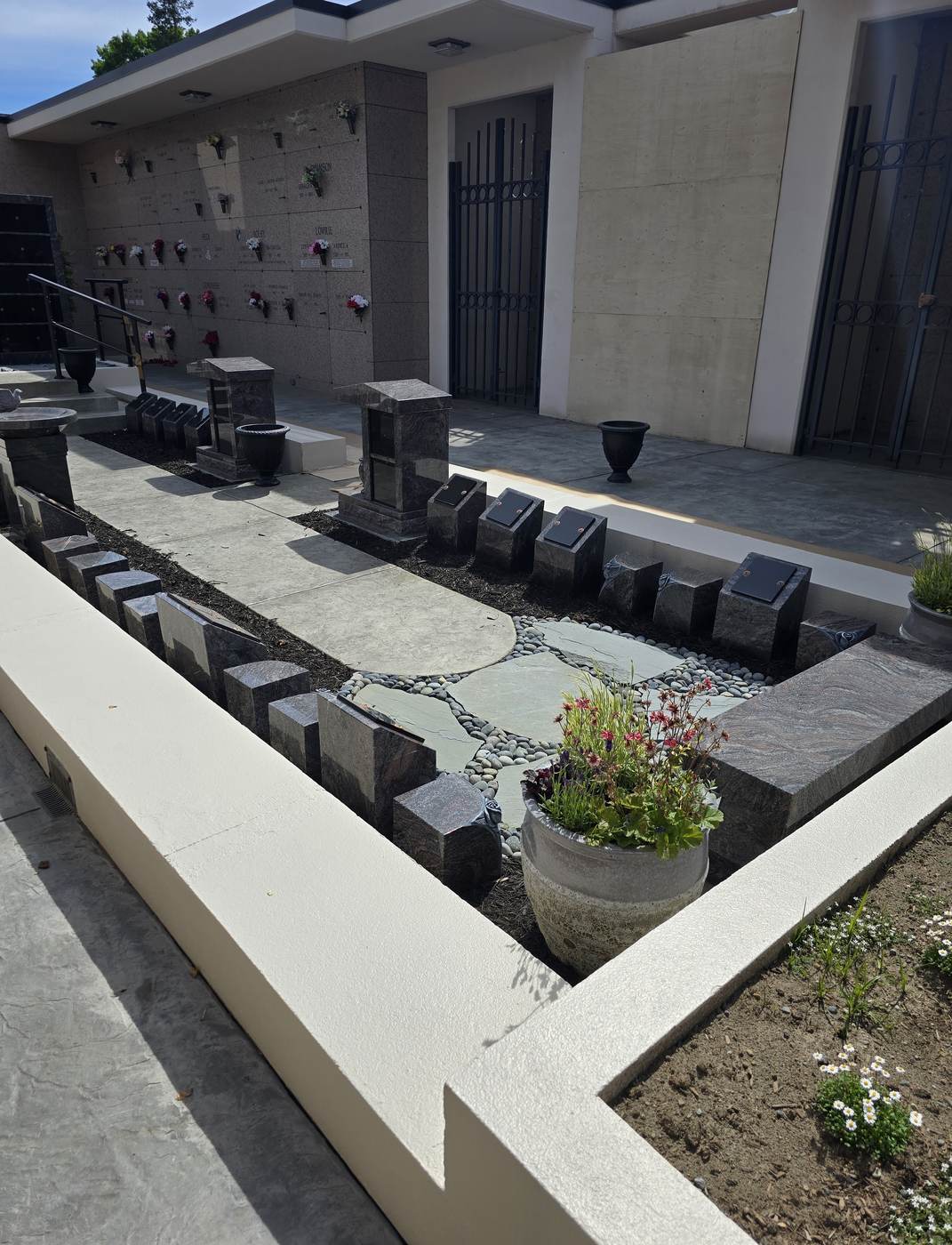The planted memorial bed along the Terrace Garden path: a row of granite cremation posts and pedestals with polished black shutters, set either side of a bluestone and river-rock path