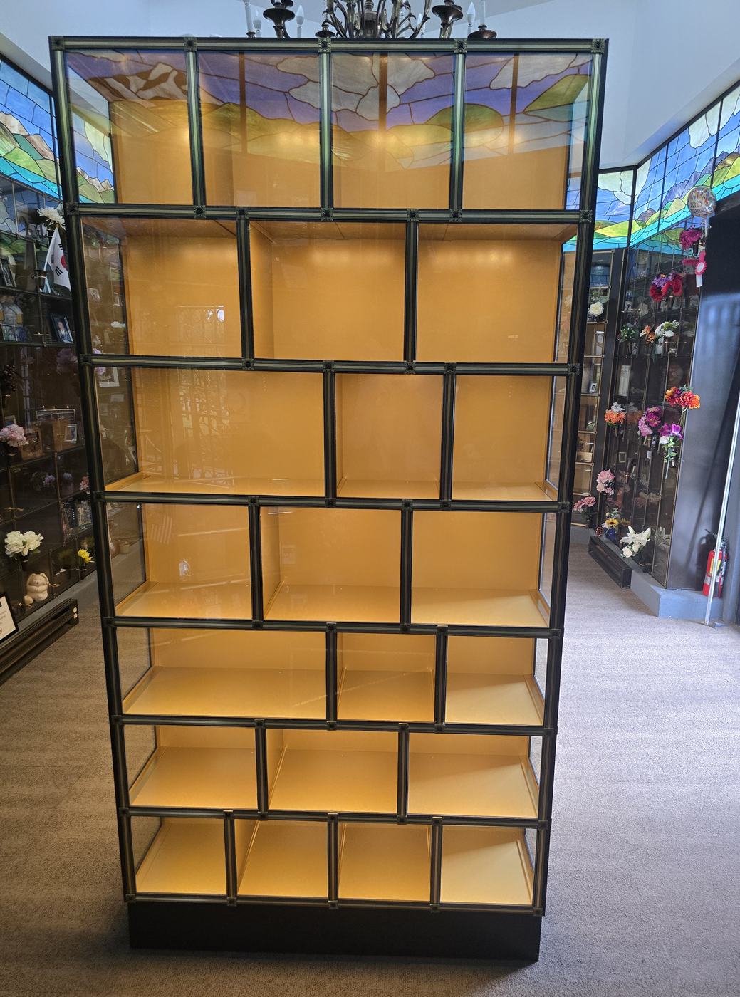 One end of the glass-front island: a full-height column of lit empty niches in bronze frames, varying in width and height, with stained glass across the top course