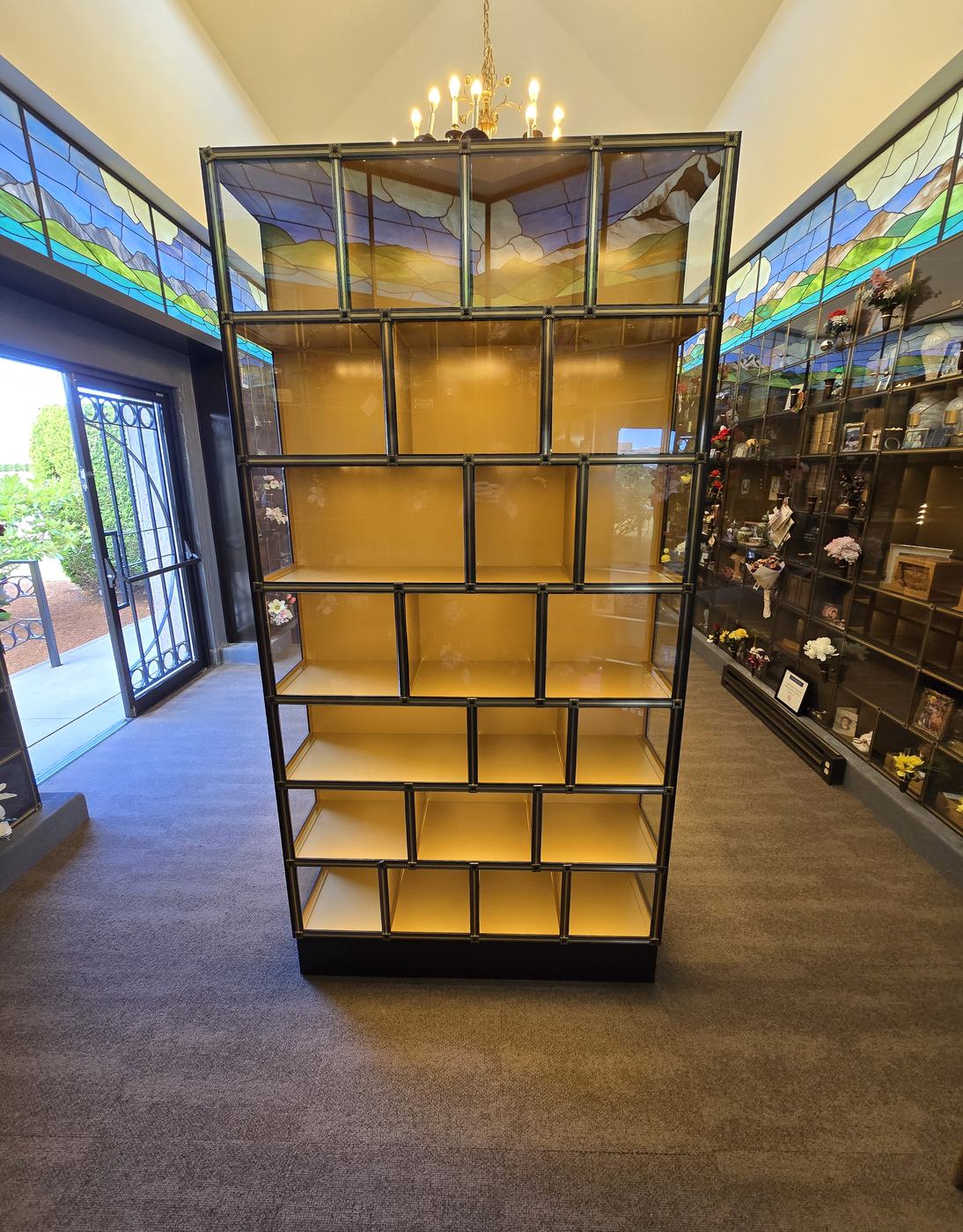 The new glass-front niche island at the Mountain View Columbarium: a tall freestanding case of lit, empty niches with bronze framing, standing under a chandelier with stained-glass panels above and a doorway to the lawn beyond