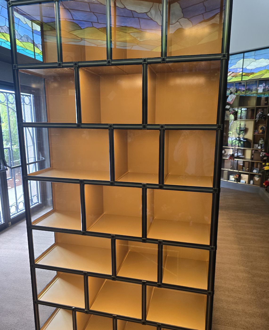 A long face of the glass-front island seen at an angle: rows of lit empty niches of several widths behind clear glass in bronze frames, with a stained-glass panel above