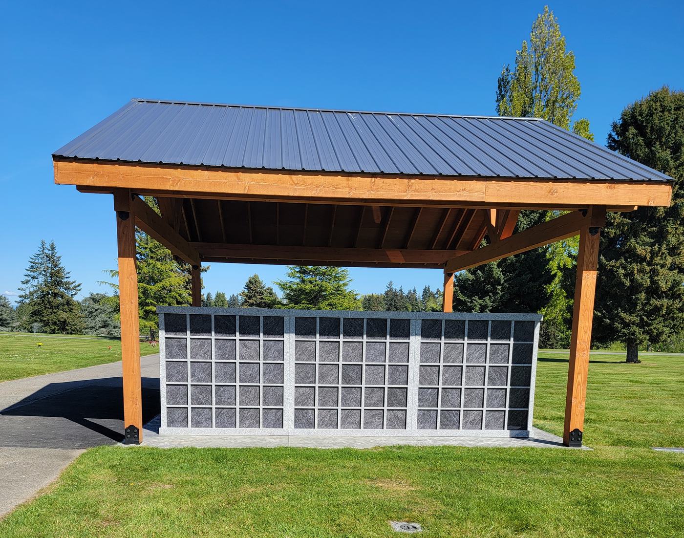 The Rock of Ages Columbarium seen from the lawn: a grey and dark granite niche wall standing under an open timber pavilion with a dark metal roof, on mown grass under a blue sky