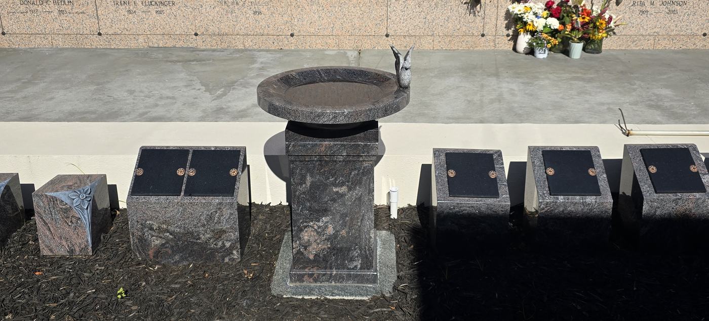 A granite birdbath on a tall pedestal standing in a bark bed, flanked by cremation posts with polished black shutters, with the mausoleum wall behind