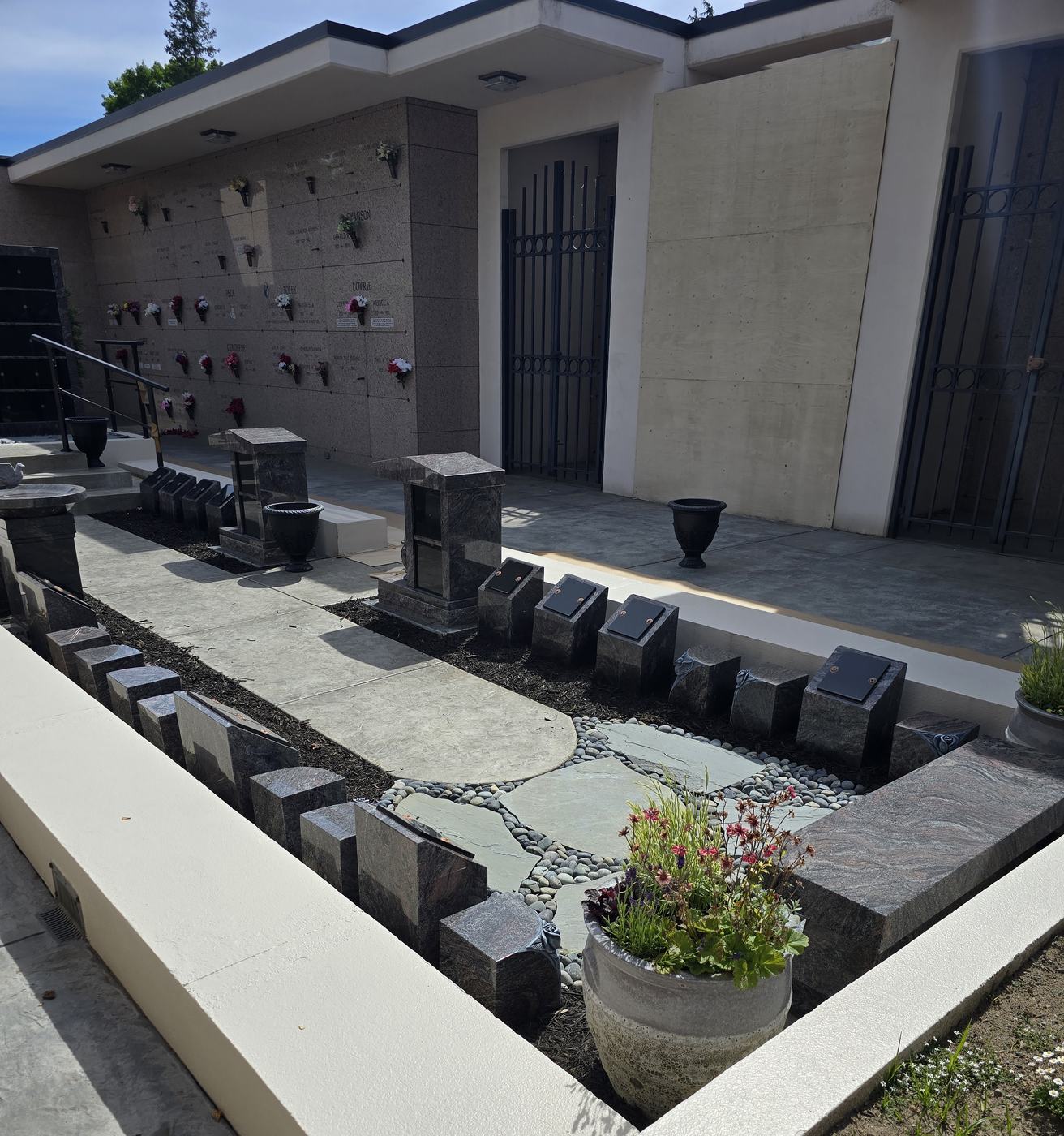 The Terrace Garden Memorial Path: a paved terrace between mausoleum wings with a bluestone and river-rock circle at its centre, rows of small granite cremation posts with polished black shutters set into dark bark beds on both sides, and a planted stone urn in the foreground