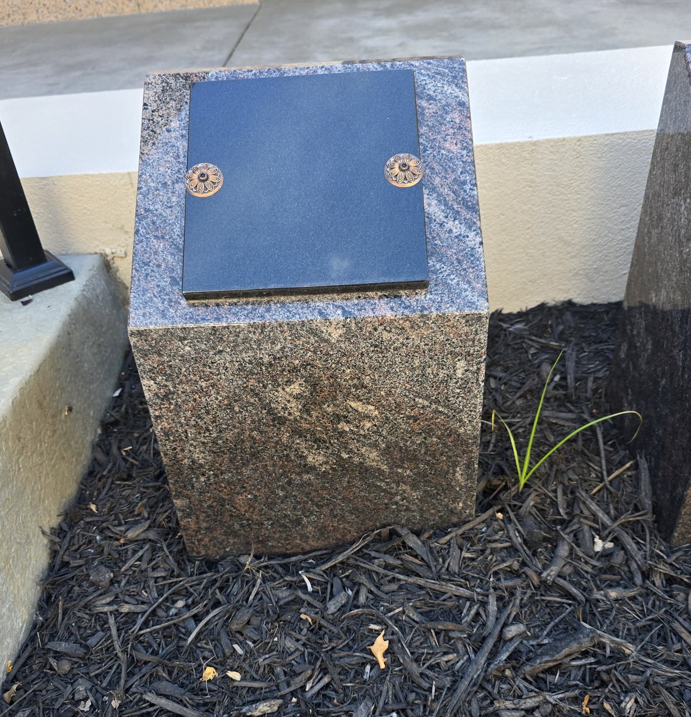 A single Paradiso cremation post in a bark bed: a small block of pink and grey granite with a polished black shutter on its sloped top, held by two bronze rosettes