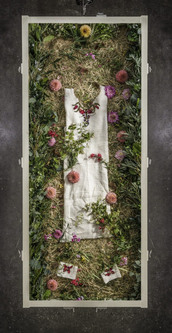 A terramation vessel from above: a person in a compostable garment on a bed of straw and greenery, dressed with flowers by the family