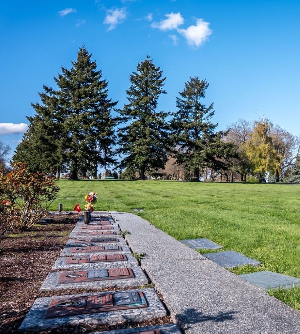 A row of bronze memorials set into a granite border along a walkway in an urn garden, with flowers in a vase, evergreens and open lawn behind