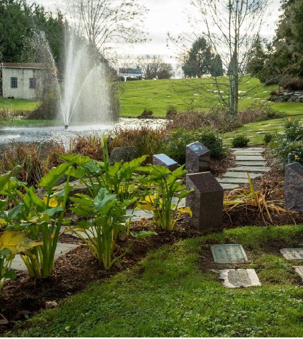 Granite cremation posts standing in a planted garden bed beside a lake fountain, with flush bronze memorials set into the grass along a flagstone path